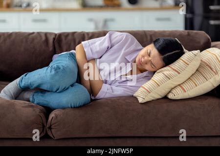 Junge Frau, die zu Hause an Bauchschmerzen leidet und auf der Couch liegt Stockfoto