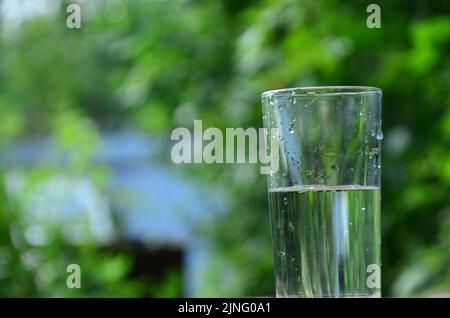 Trinken Sie Wasser, das im Freien über Sonnenlicht und natürlichem grünen Hintergrund auf dem weißen Tisch in Glas fließt Stockfoto