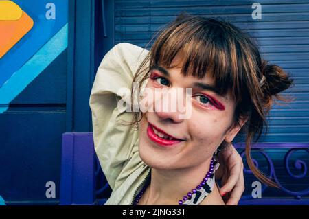 Horizontales Nahaufnahme-Porträt einer glücklichen Transgender-latin-jungen Frau mit Make-up, die auf die Kamera schaut und lächelt und mit Blau auf der Straße posiert Stockfoto