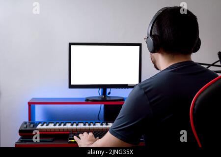 Rückansicht eines jungen kaukasischen Mannes mit Kopfhörern, der mit einer Hand auf der Tastatur den Computermonitor beobachtete, in seinem Heimmusik-Studio, Technologie und Stockfoto