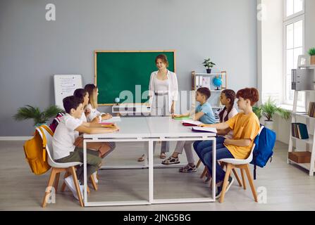 Glückliche Schüler sitzen im Klassenzimmer am Tisch, lesen Bücher und sprechen mit dem Lehrer Stockfoto