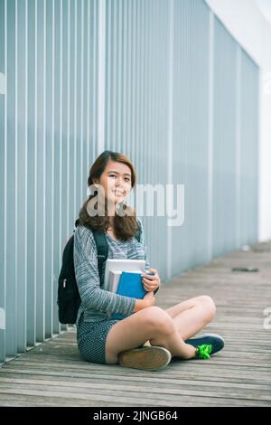 Fröhliches vietnamesisches Schulmädchen mit Lehrbüchern, die auf dem Boden sitzen Stockfoto