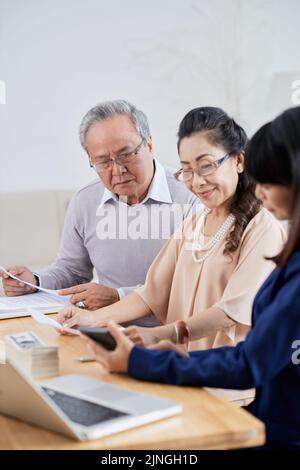 Immobilienkauf: Senior Asian Paar sitzt im modernen Büro und zahlt für ihre neue Wohnung an Pretty makler nach erfolgreichem Abschluss der Sitzung Stockfoto