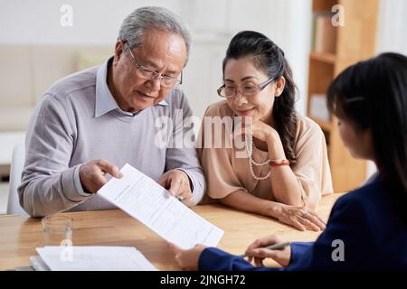 Konzentrierter Senior Mann und seine hübsche Frau studieren Bedingungen des Immobilienkaufvertrags, während mit makler, Interieur des modernen Büros auf dem Hintergrund treffen Stockfoto