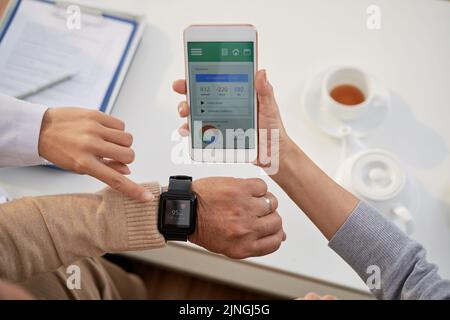 Ein junger Arzt mit weißem Mantel erklärt älteren Patienten, wie sie die mobile Anwendung zur Kontrolle der Herzfrequenz nutzen können, während sie sich im Büro beraten Stockfoto