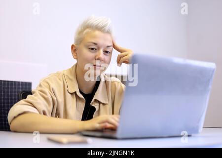 Lächelndes Mädchen mit kurzen blonden Haaren sitzt am Bürotisch mit Laptop. Tomboy Lifestyle, Konzept der Inspiration bei der Arbeit und Kreativität Stockfoto