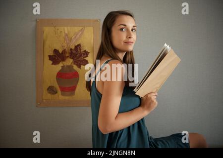Porträt einer schönen jungen Frau mit offenem Buch, Bild mit Herbstblättern an der Wand hinter ihr Stockfoto