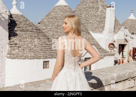 Rückansicht der jungen glücklichen Frau Braut, die in der Nähe von konischen Dächern von Trullo im sonnigen Italien steht und die Hand auf die Taille posiert. Stockfoto