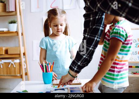 Zwei niedliche kleine Mädchen, die an einem weißen Tisch stehen und Brettspiele im hellen Klassenzimmer spielen. Lehrer hilft Kindern. Stockfoto