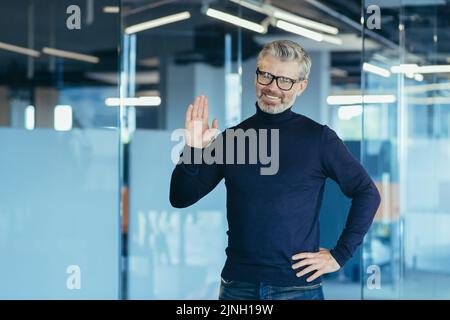 Porträt eines erfolgreichen Finanziers im modernen Büro, eines Mannes, der die Kamera anschaut und lächelnd die Hand hochhält, eines erfahrenen, grauhaarigen Geschäftsmanns mit Bart und Brille, eines Investors Stockfoto