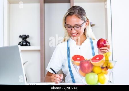 Lächelnde Ernährungsberaterin in ihrem Büro hält sie Obst in der Hand und zeigt gesundes Gemüse und Obst, Gesundheits- und Ernährungskonzept. Ernährungsberaterin Stockfoto