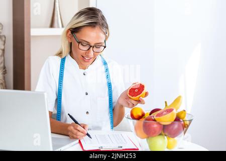 Porträt einer jungen lächelnden Ernährungsberaterin im Sprechzimmer. Ernährungsberater mit gesundem Obst, Saft und Maßband. Diätassistent arbeitet Stockfoto