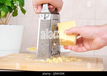 Ein Mann reibt Käse auf einer Metallreibe zum Einfrieren und zum weiteren Kochen von Pizza, Pasta. Stockfoto