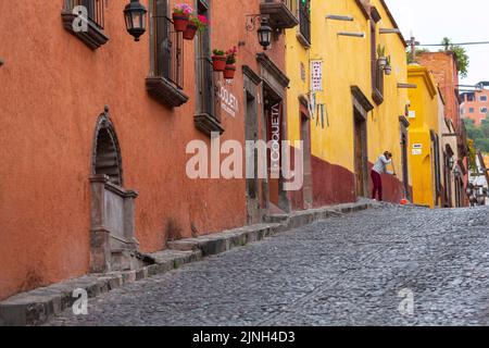 Eine Frau geht über den Gang in der Calle Cuadrante, die von farbenfrohen Gebäuden im spanischen Kolonialstil gesäumt ist, im historischen Stadtzentrum von San Miguel de Allende, Mexiko. Stockfoto