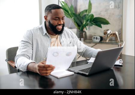 Fokussierter erfolgreicher, positiv-afroamerikanischer, vielbeschäftigter Mann, Freiberufler oder Büroangestellter, der Dokumente arbeitet, an einem Schreibtisch im modernen Büro sitzt, an einem Projekt arbeitet, verwendet Laptop, studiert Graphen, lächelt Stockfoto