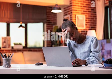 Erschöpfte Außendienstmitarbeiterin, die Kopfschmerzen hatte, junge gestresste Freiberuflerin, die am Laptop arbeitete. Unglücklicher afroamerikanischer Mitarbeiter am Arbeitsplatz im Heimbüro mit schönem warmen Abendlicht Stockfoto