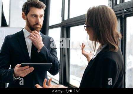 Eleganter kaukasischer Mann in einem formellen Anzug, kommuniziert mit seiner Kollegin, sie stehen im Büro, diskutieren Arbeitsfragen, Finanzberichte, Unternehmensgewinne, entwickeln Ideen Stockfoto