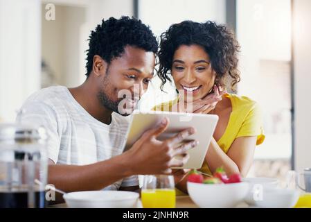 Schauen Sie sich auch das an, es ist unglaublich. Ein junges Paar, das ein digitales Tablet zusammen benutzt, während es zu Hause frühstückt. Stockfoto