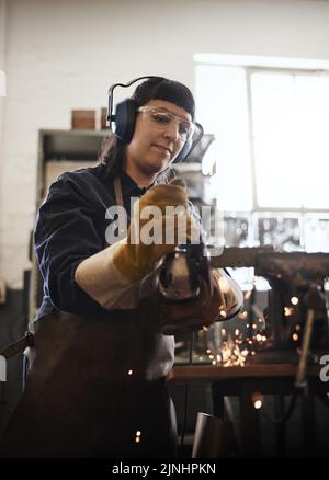 Eine attraktive junge Kunsthandwerkerin, die in ihrer Werkstatt einen Winkelschleifer verwendet. Stockfoto