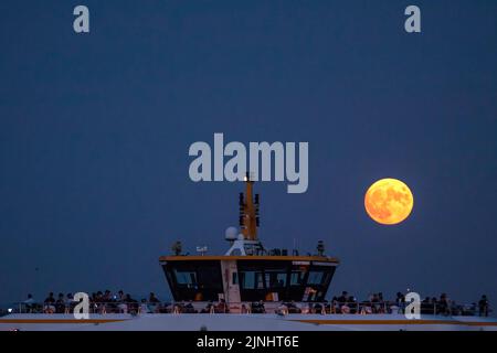 Istanbul, Türkei. 11. August 2022. Mit dem Vollmond am Himmel wurden Passagiere auf der Terrasse der City Lines Fähre gesehen. Boote, die durch den Bosporus und die Stadtlinien-Fähren fuhren, wurden in Istanbul gesehen, wobei der Vollmond am Himmel in den Abendstunden erschien. Kredit: SOPA Images Limited/Alamy Live Nachrichten Stockfoto