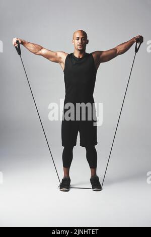 Haben Sie keine Angst, sich auszudehnen. Studioporträt eines athletischen jungen Mannes, der mit einem Widerstandsband vor grauem Hintergrund arbeitet. Stockfoto