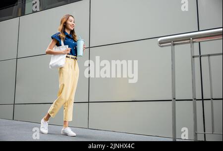 Asiatische schöne Geschäftsfrau zuversichtlich lächelnd mit Stofftasche hält Stahl Thermoskannen Becher Wasser Glas sie zu Fuß im Freien auf der Straße in der Nähe Modus Stockfoto