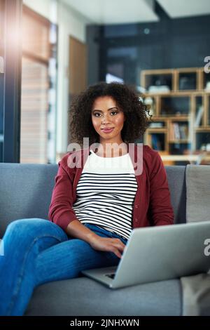 Mein neuer Blog ist im Aufbau. Porträt einer jungen Frau, die einen Laptop benutzt, während sie sich zu Hause auf dem Sofa entspannt. Stockfoto