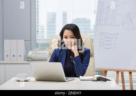 Portrait eines attraktiven asiatischen Unternehmers, der am Schreibtisch sitzt und mit Hilfe eines Laptops an einem vielversprechenden Projekt arbeitet, Büroinnenraum im Hintergrund Stockfoto
