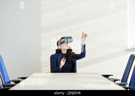 Junge Designerin mit langem lockigen Haar, die ein VR-Headset verwendet, während sie in einem geräumigen Sitzungssaal sitzt und an einem neuen Projekt zur Visualisierung von 3D arbeitet Stockfoto