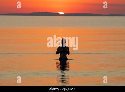 Portobello, Edinburgh, Schottland, Großbritannien. 12.. August 2022. Atemberaubender Sonnenaufgang, um eine sehr heiße Woche mit überdurchschnittlichen Temperaturen für Schottland zu beenden, Temperatur heute Morgen im Morgengrauen 13 Grad Celsius für diejenigen, die am Firth of Forth Seaside Sport treiben wollen, wobei haar später an der Ostküste erwartet wird. Im Bild: Diese junge Frau genießt ein kaltes Wasserbad in der Morgendämmerung, gerade als die Sonne beginnt, über den Horizont zu blicken. Quelle: Arch White/alamy Live News. Stockfoto