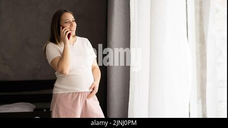 Schwangere, die im Wohnzimmer zu Hause mit warmem Licht, Transparent und leerem Raum auf dem Mobiltelefon spricht Stockfoto