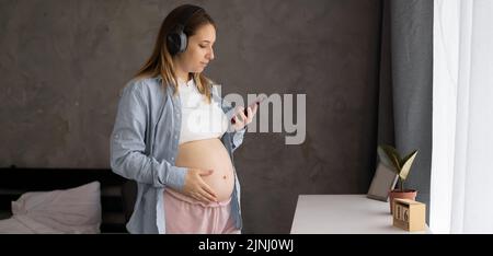 Junge Schwangere hören Musik über Kopfhörer mit dem Smartphone, Schwangere entspannen sich im Wohnzimmer. Schwangerschaft, Technolo Stockfoto