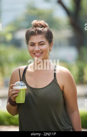 Porträt einer attraktiven jungen Frau, die mit einem breiten Lächeln auf die Kamera blickt und einen Plastikbecher mit Smoothie in der Hand hält, während sie den Sommertag im öffentlichen Park genießt Stockfoto