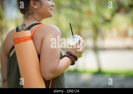 Lächelnde, übergewichtige Frau, die wegschaut und die malerische Aussicht auf den öffentlichen Park genießt, während sie sich nach einem produktiven Fitnesstraining mit Smoothie erfrischt Stockfoto