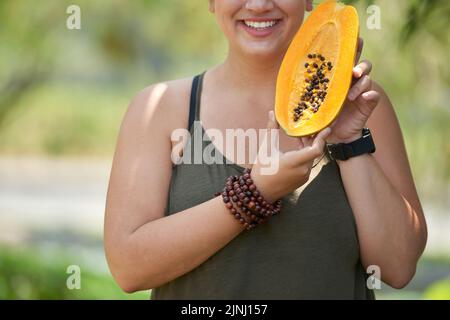Nicht erkennbare junge Frau mit einem charmanten Lächeln, die in einem grünen, von Sonnenstrahlen beleuchteten Park steht und reife Papaya in den Händen hält Stockfoto