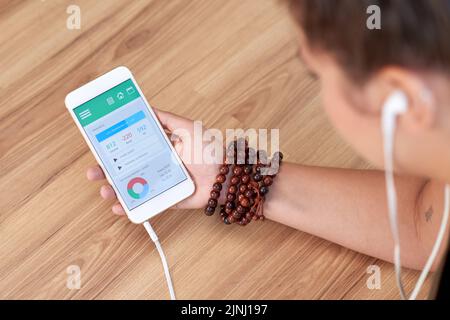 Nahaufnahme einer nicht erkennbaren Frau, die am Holztisch sitzt und die Gesundheitsüberwachungsanwendung auf dem Smartphone verwendet Stockfoto