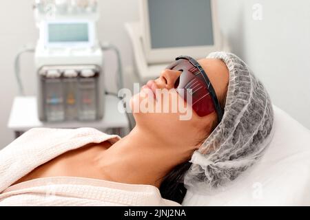 Das Porträt einer jungen Frau in einer Schutzbrille liegt auf der Couche im Schönheitssalon. Konzept der professionellen Kosmetologie und plastischen Chirurgie. Stockfoto