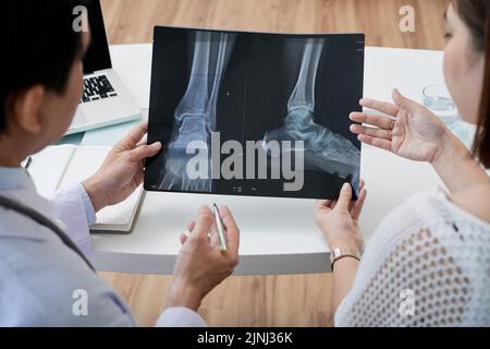 Über die Schulter eines Chirurgen mittleren Alters, der einer Patientin während eines Termins eine Fußröntgenaufnahme zeigt Stockfoto