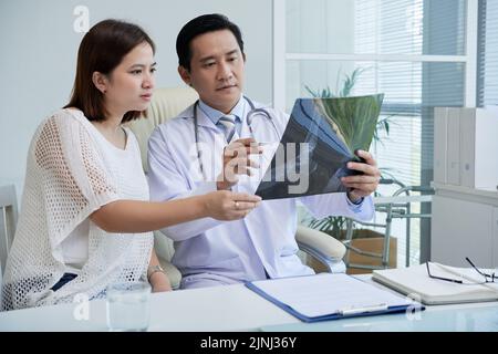 Ein hübscher Chirurg mittleren Alters mit weißem Mantel sitzt am Schreibtisch und zeigt dem hübschen Patienten ein Röntgenbild, während er einen Termin im modernen Büro hat Stockfoto