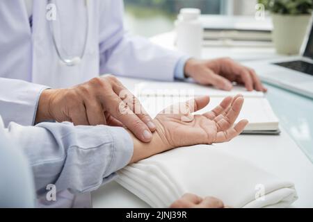 Nahaufnahme eines nicht erkennbaren Kardiologen mit weißem Mantel, der am Schreibtisch sitzt und die Pulsfrequenz einer weiblichen Patientin misst Stockfoto