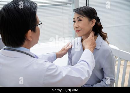Selbstbewusster Arzt mittleren Alters, der Brillen trägt und Drüsen von älteren Patienten untersucht, während er einen Termin im modernen Büro hat Stockfoto
