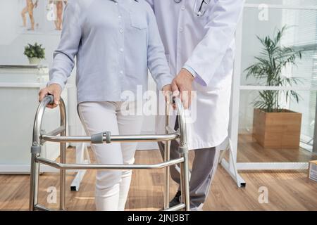 Nahaufnahme eines nicht erkennbaren Physiotherapeuten, der dem Patienten erklärt, wie er einen Vorderradspaziergänger benutzt, Inneneinrichtung eines modernen Büros im Hintergrund Stockfoto