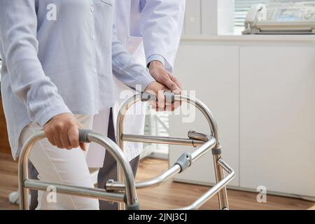 Nicht erkennbare Physiotherapeutin mit weißem Mantel neben der Patientin stehend, während sie ihr dabei half, den Frontrollerwagen richtig zu verwenden, die Inneneinrichtung des modernen Büros auf dem Hintergrund Stockfoto