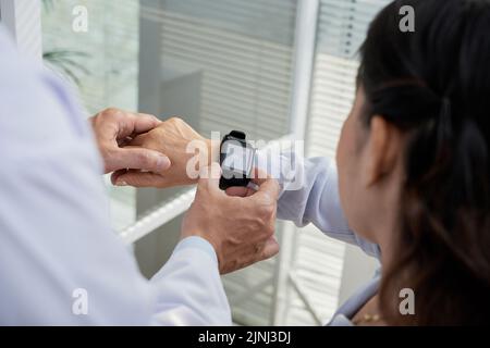 Über der Schulter zeigt ein freundlicher Therapeut seinem älteren Patienten, wie er mithilfe der Smartwatch und Nahaufnahme die Herzfrequenz überprüfen kann Stockfoto