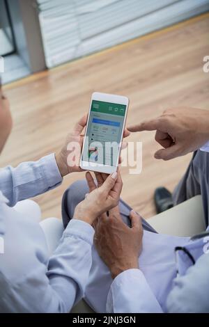Nahaufnahme eines nicht erkennbaren Arztes mit weißem Mantel, der auf dem Sofa eines modernen Büros sitzt und der weiblichen Patientin erklärt, wie sie die App zur Gesundheitsüberwachung auf dem Mobiltelefon nutzt Stockfoto