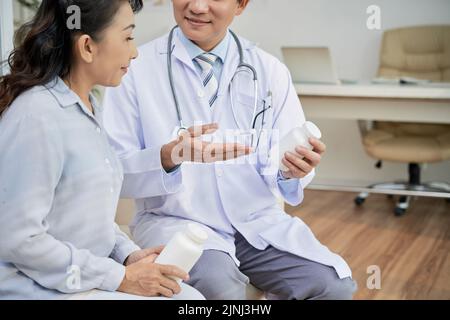 Profilansicht einer älteren asiatischen Patientin, die ihrem Arzt mit Interesse zuhört, während er ihr Anweisungen gibt, wie man Pillen richtig einnimmt, Inneneinrichtung eines modernen Büros im Hintergrund Stockfoto