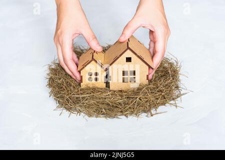 Hände legen zwei Miniatur-Holzhäuser in ein Heunest isoliert auf neutralem Hintergrund. Familienhauskonzept. Stockfoto