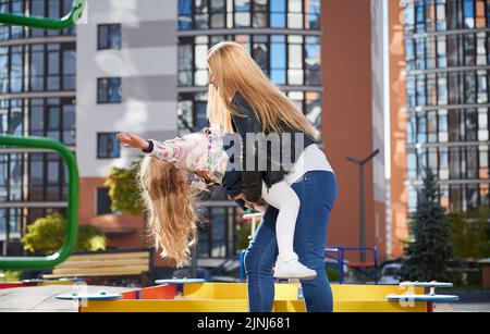 Weiblich hält liebenswert Tochter auf dem Kopf auf Spielplatz. Mädchen, die Spaß mit attraktiven blonden Mutter im modernen Innenhof der Stadt Wohnhochhäuser. Konzept von Familie und Kindheit. Stockfoto