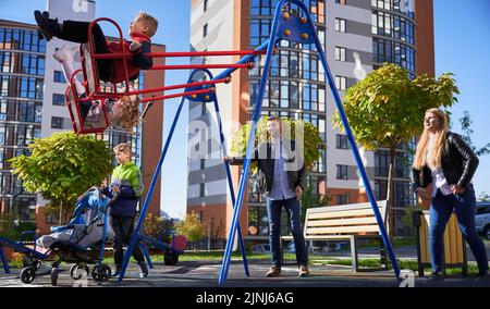 Glückliche Familie - Vater, Mutter und Kinder, die gemeinsam auf dem Spielplatz Spaß haben. Eltern schwingen Tochter und Sohn, während ein anderes Kind mit Kinderwagen steht. Moderne Wohngebäude im Hintergrund. Stockfoto