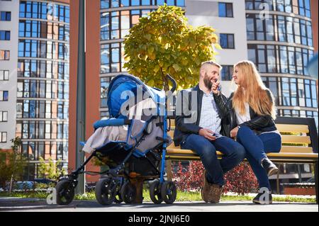 Glückliche Familie, die zusammen mit einem nicht erkennbaren Baby in einem blauen Kinderwagen im modernen Innenhof eines Wohnhochgebäudes der Stadt sitzt. Lächelnde Frau mit den Schlüsseln aus der neuen Wohnung. Stockfoto